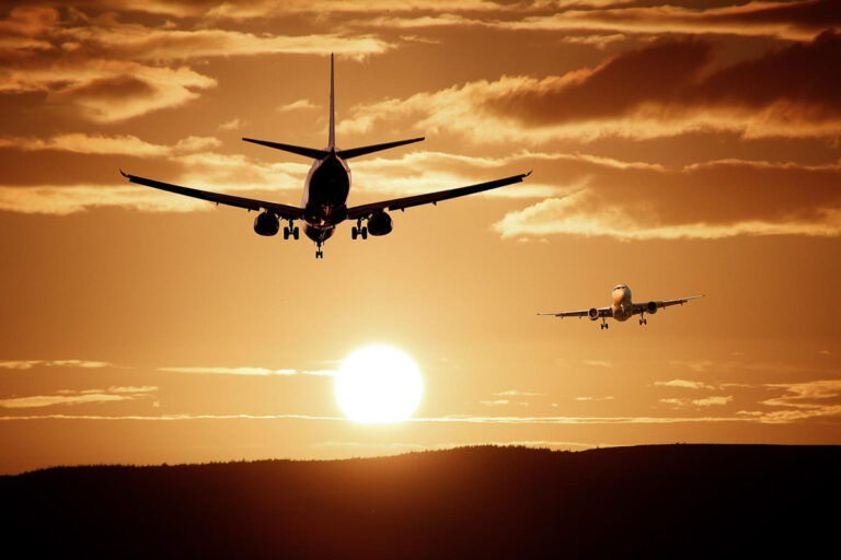 Two airplanes are flying in the sky during sunset, with the sun low on the horizon and clouds scattered above.