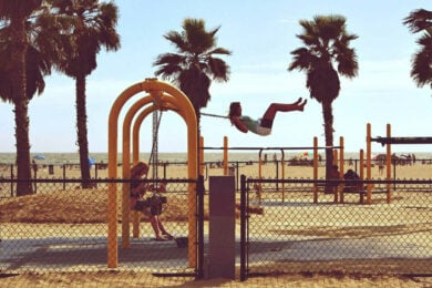 Two children swing on yellow swings at a beachside playground, with palm trees, sand, and the ocean visible in the background.