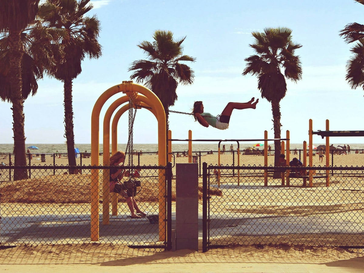 Two children swing on yellow swings at a beachside playground, with palm trees, sand, and the ocean visible in the background.