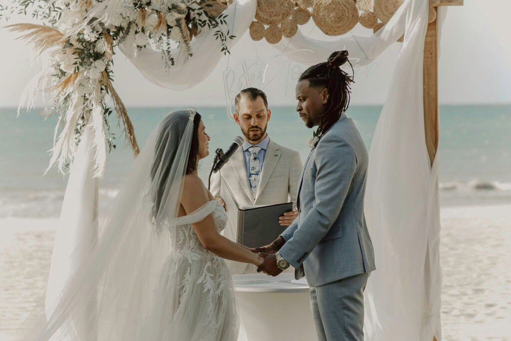 A bride and groom hold hands at a beachside wedding ceremony, standing under a decorated arch with an officiant reading from a folder.