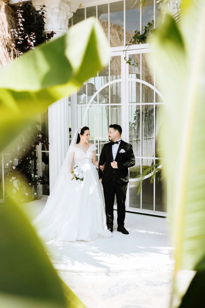 A bride and groom stand together in front of large glass doors, surrounded by greenery, with sunlight filtering through leaves in the foreground.