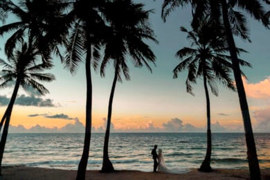 A couple stands on a beach at sunset, framed by tall palm trees, with the ocean and a colorful sky in the background.