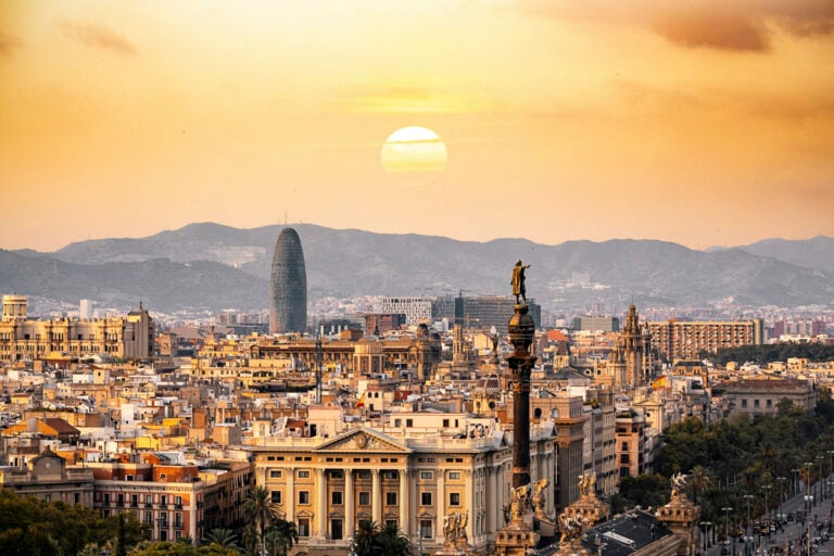 Aerial view of Barcelona at sunset with the sun low in the sky, city buildings, the Columbus Monument, and mountains in the background.