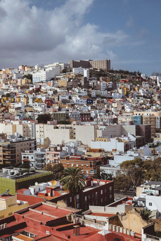 A densely packed hillside cityscape with colorful buildings, a large apartment complex at the top, and partly cloudy skies overhead.