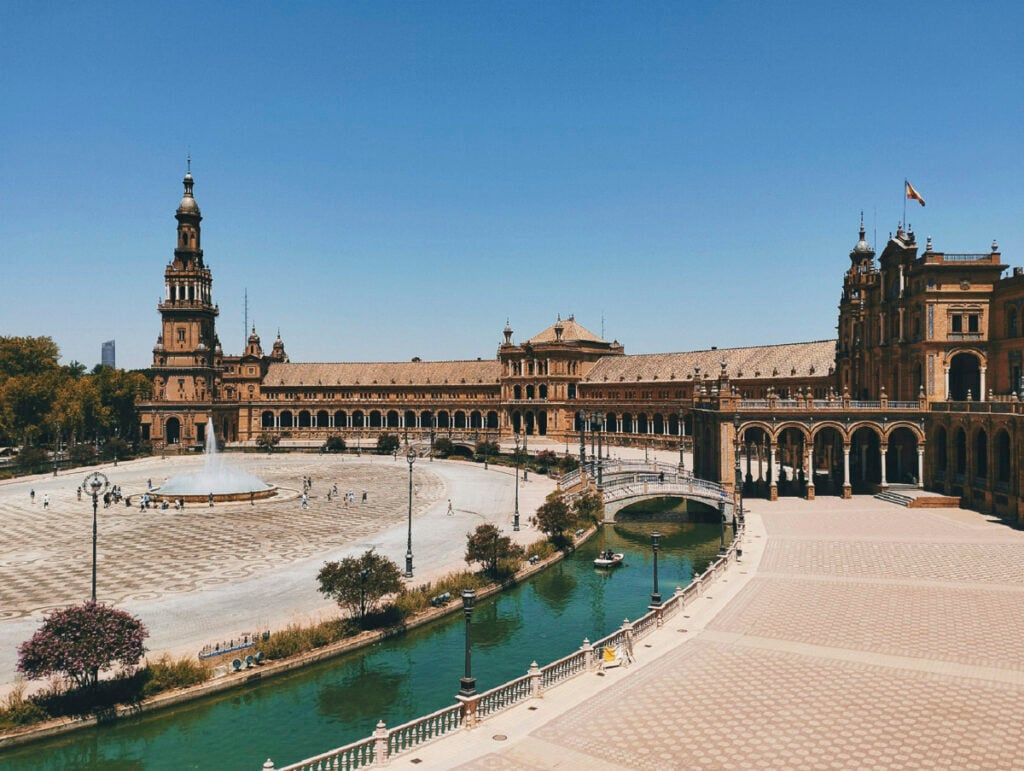 A wide view of Plaza de Espa&ntilde;a in Seville, Spain, showing its semicircular building, central fountain, canal, and people walking in the square under a clear blue sky.