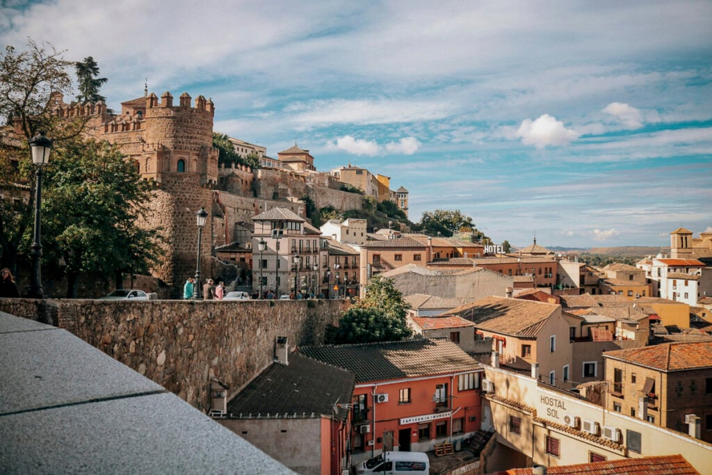 View of historic stone buildings and a fortified wall overlooking a cluster of rooftops in Toledo, Spain, under a partly cloudy sky.