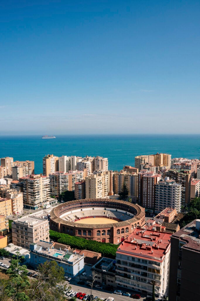 Aerial view of a coastal city with a bullring in the center, surrounded by apartment buildings, and the sea visible in the background under a clear blue sky.