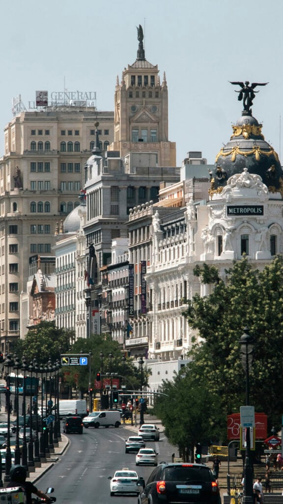 A busy city street with cars and ornate historic buildings, including the Metropolis Building with its dome and statue, under a clear sky.