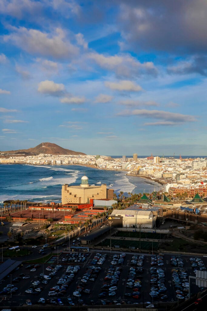 A coastal cityscape with a beach, a prominent building with a dome, a parking lot in the foreground, and mountains in the background under a partly cloudy sky.