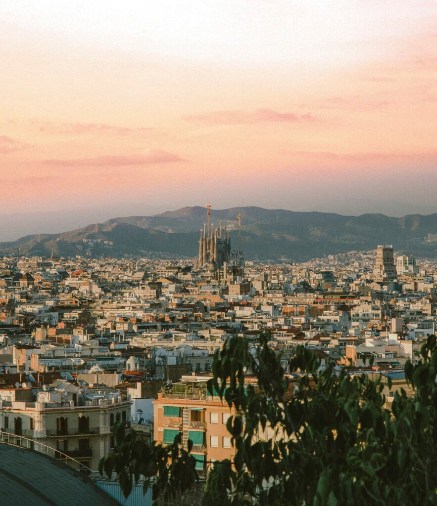 A wide view of Barcelona at sunset, with the Sagrada Familia basilica under construction in the center and mountains in the background.
