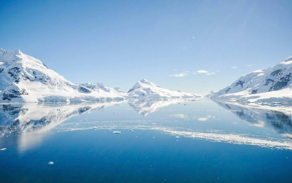 Snow-covered mountains reflected in a calm, icy body of water under a clear blue sky.