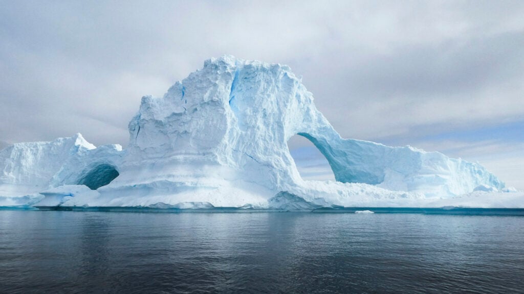 Large iceberg with arch-shaped openings floating in calm, dark water under a cloudy sky.