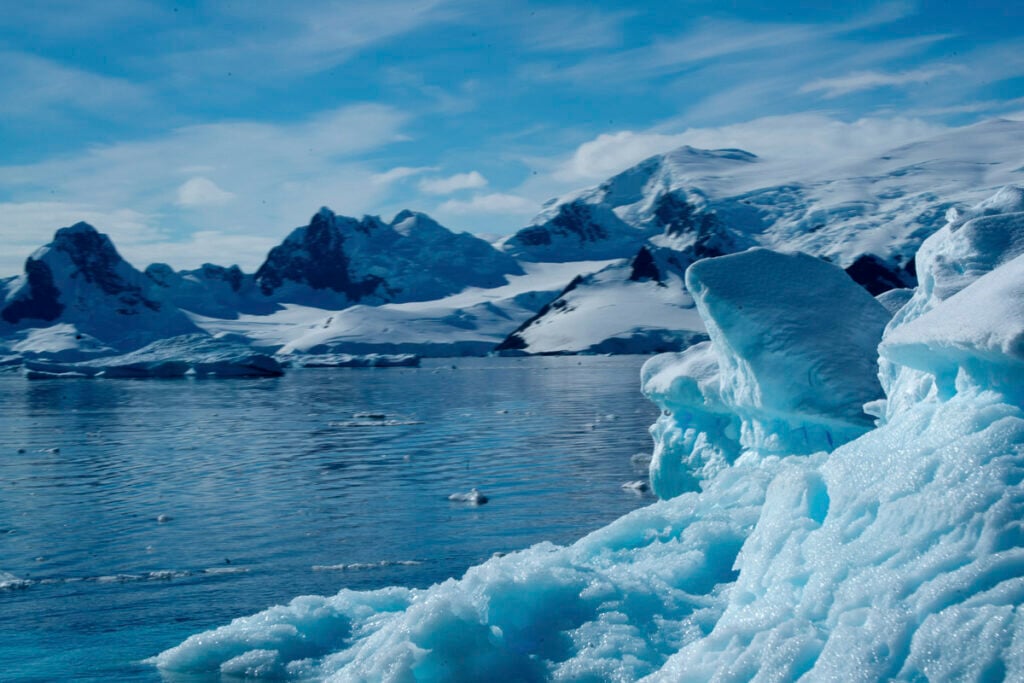 Snow-covered mountains and glaciers rise above a calm body of water under a blue sky with scattered clouds.