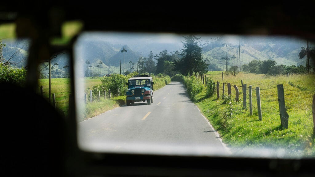 A blue and white vintage jeep drives down a rural road lined with grass and fence posts, with mountains visible in the background.