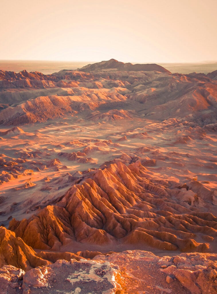 A vast desert landscape with rugged, sunlit mountains and ridges under a clear sky, viewed from an elevated vantage point.