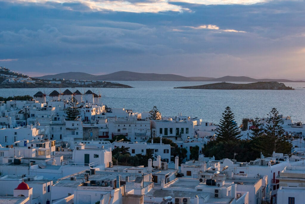 White buildings with flat roofs overlook the sea at sunset, with several windmills and distant islands under a partly cloudy sky.