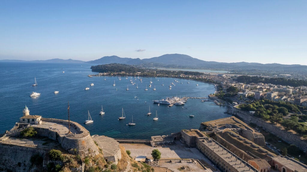 Aerial view of a coastal town with a harbor filled with boats, historic fortifications in the foreground, and mountains in the background under a clear sky.