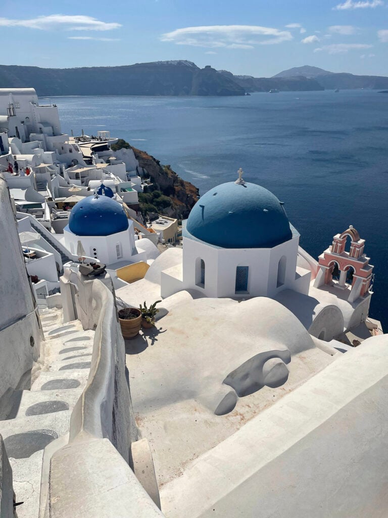 White buildings with blue domes overlook the Aegean Sea in Santorini, Greece, with cliffs and distant islands visible under a partly cloudy sky.