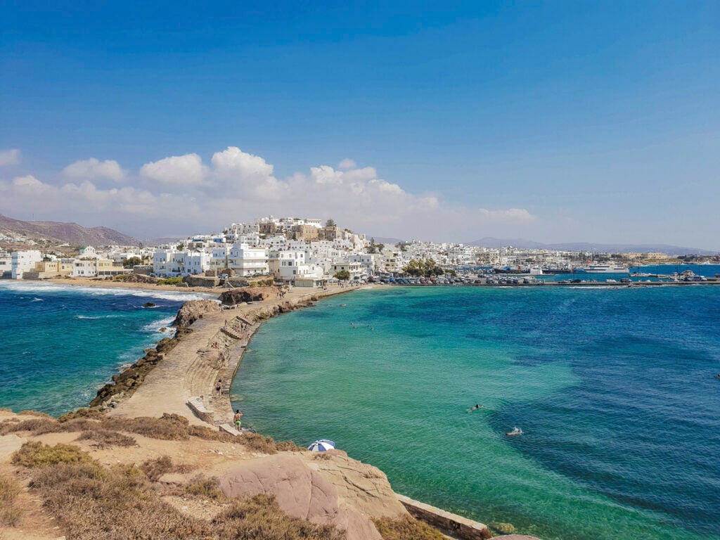 Coastal view of a town with white buildings on a hill, surrounded by turquoise sea, a stone walkway, and people swimming in the water under a clear blue sky.