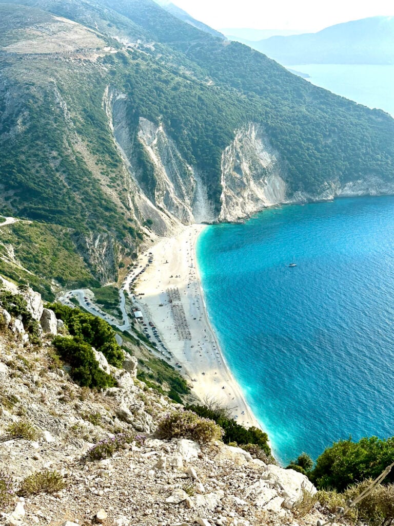 Aerial view of a sandy beach with turquoise water, backed by steep cliffs and green hills, with sunbeds and umbrellas arranged along the shoreline.