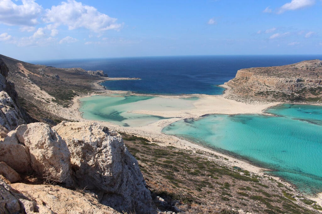 A coastal landscape with clear turquoise lagoons, sandy beaches, and rocky hills under a mostly clear sky with scattered clouds.