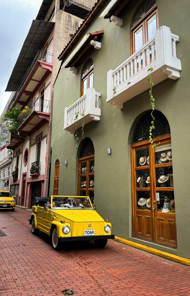A yellow convertible with two passengers drives on a brick street past a green building with arched windows, balconies, and hats displayed inside.