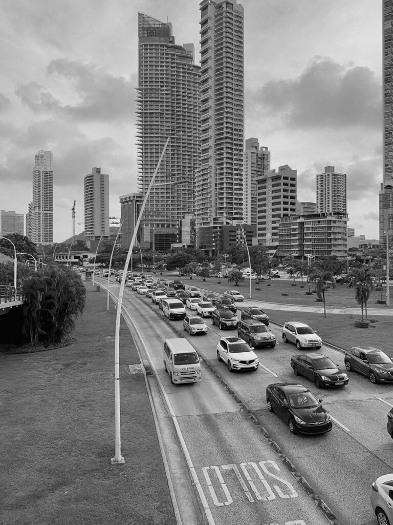 Black and white photo of a busy city street with multiple lanes of cars in traffic, surrounded by tall modern buildings and cloudy skies.
