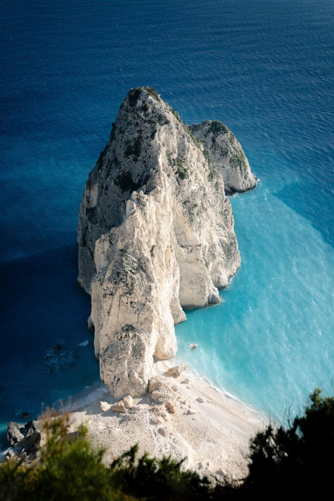 A large rocky formation rises from turquoise blue water next to a sandy beach, viewed from above with green foliage in the foreground.