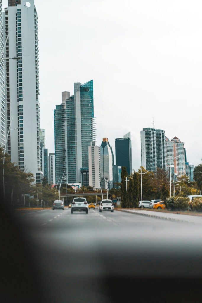 Cars drive on a city street lined with tall modern skyscrapers and trees, under an overcast sky.