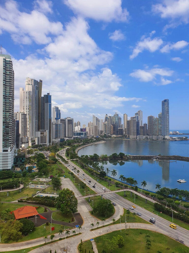 A cityscape of modern high-rise buildings lines a curved waterfront under a partly cloudy sky, with a park and busy roads in the foreground.
