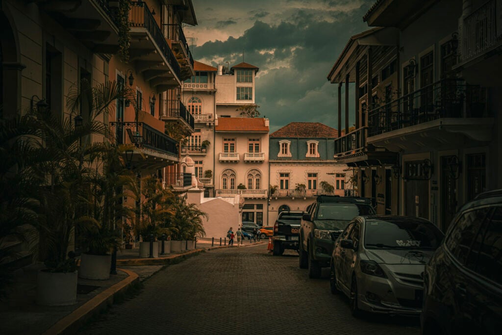 A narrow cobblestone street lined with parked cars, potted plants, and pastel-colored buildings under a cloudy sky.