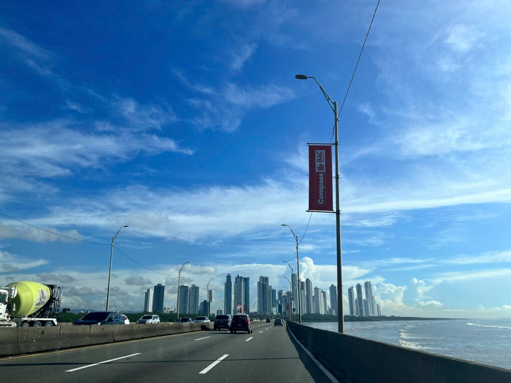 A wide highway runs along the coast with skyscrapers in the distance under a blue sky; a street lamp and a red sign are visible in the foreground.