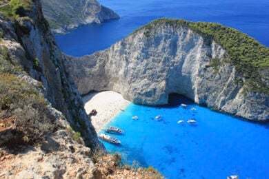 Aerial view of a small sandy beach surrounded by steep cliffs and clear blue water with several boats anchored near the shore.