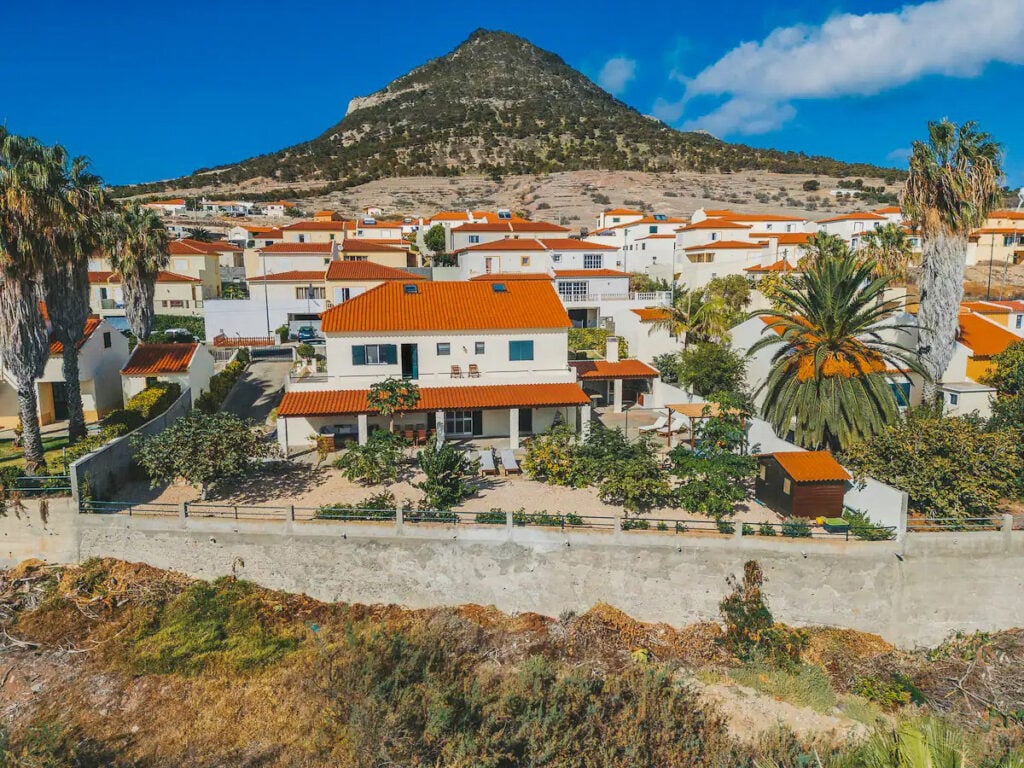 A residential neighborhood with white houses and red tile roofs, palm trees, and a mountain in the background under a clear blue sky.
