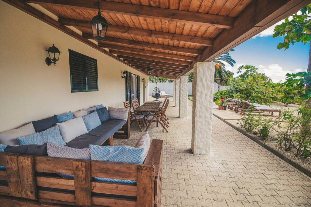 Covered patio with wooden ceiling, outdoor seating area with cushions, dining table with chairs, and view of greenery and sky.