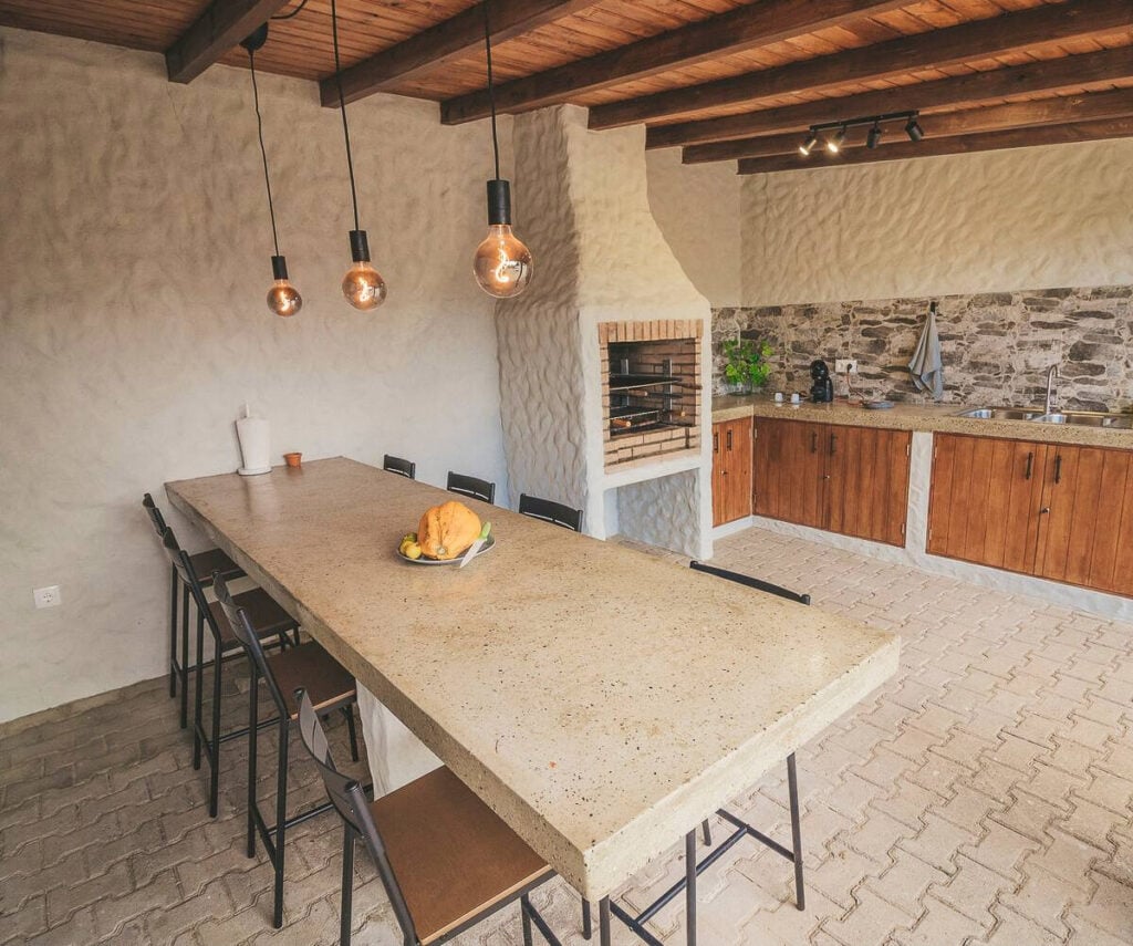 Modern indoor kitchen with a long concrete dining table, black chairs, hanging lights, built-in grill, and wooden cabinets against a stone-tiled wall.