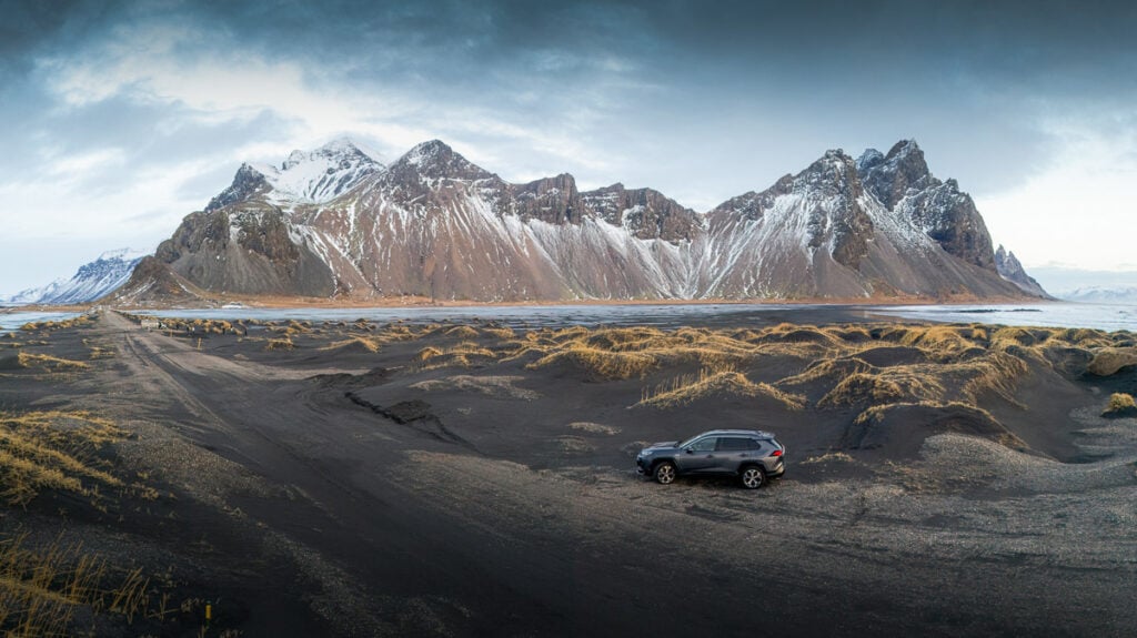 A black SUV is parked on a sandy, black landscape with snow-capped mountains in the background under a cloudy sky.