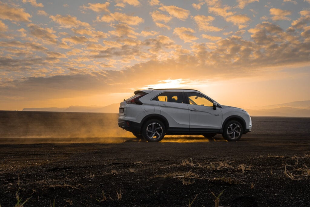 A white SUV drives on a dirt road at sunset, with dust rising around its wheels and clouds visible in the sky.