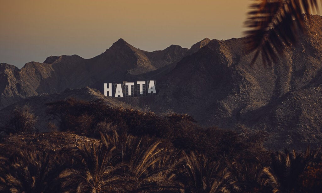 A large white sign reading “HATTA” stands on a rocky hillside, with palm trees in the foreground and rugged mountains in the background at sunset.