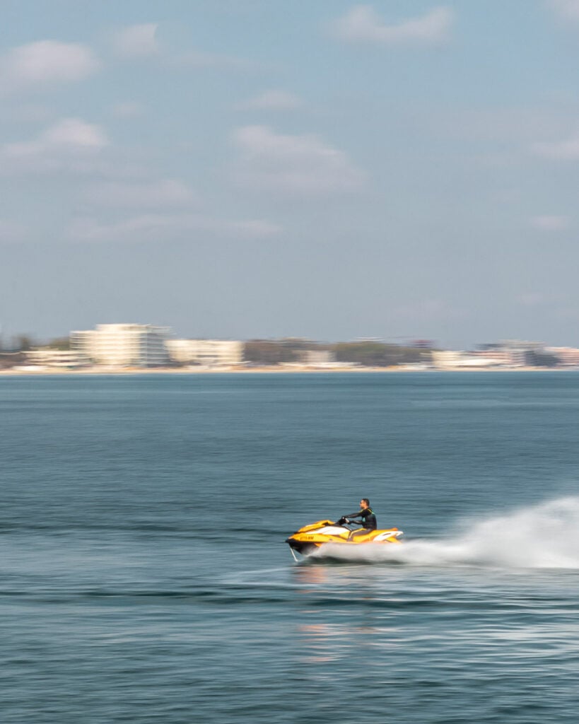 Person riding a yellow jet ski across calm blue water, with a blurred city shoreline and buildings in the background under a partly cloudy sky.