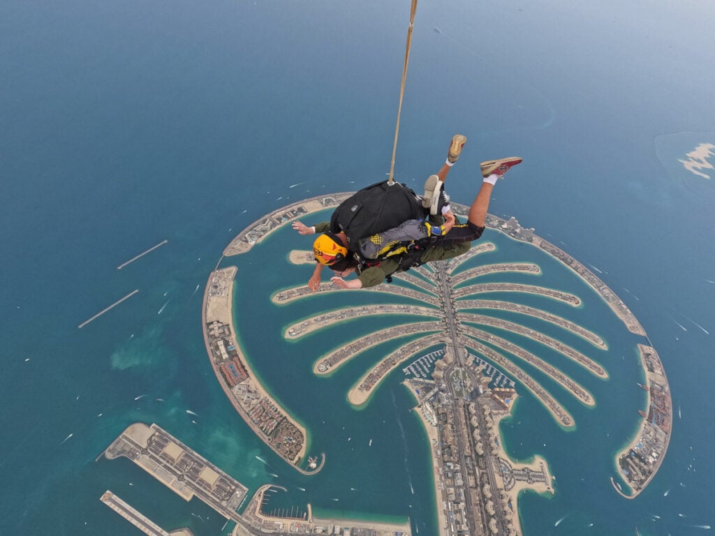 A person skydiving over the Palm Jumeirah, an artificial island shaped like a palm tree, surrounded by blue water in Dubai.