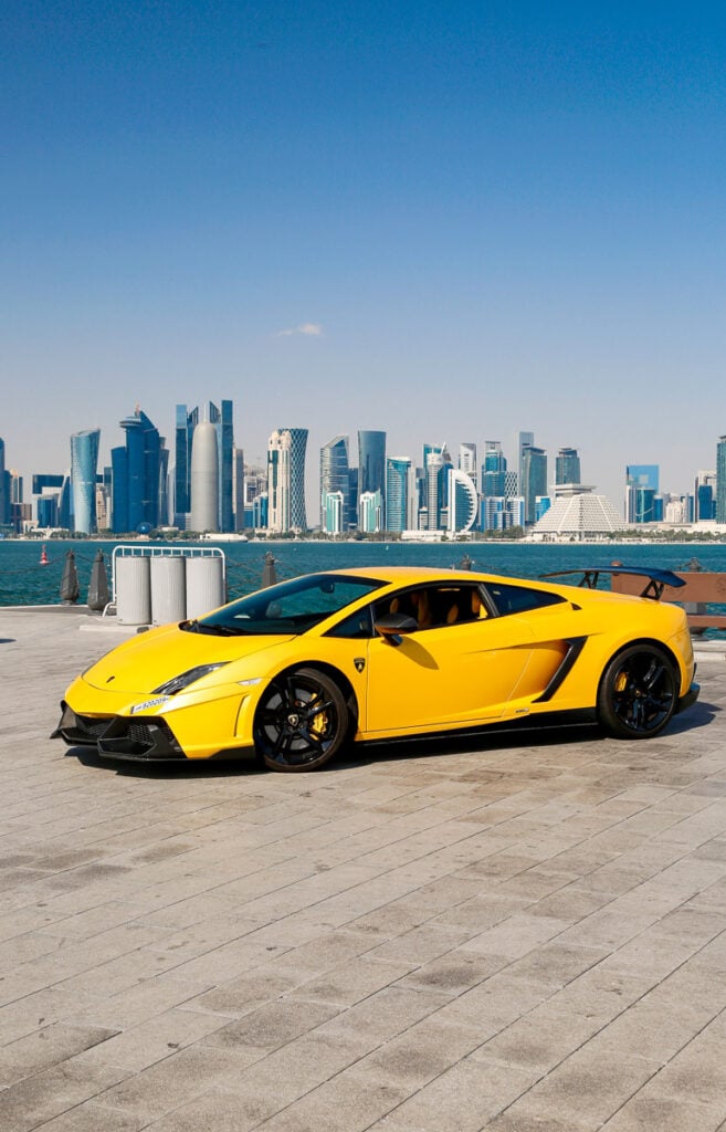 A yellow sports car is parked on a paved area near water, with a modern city skyline in the background under a clear blue sky.