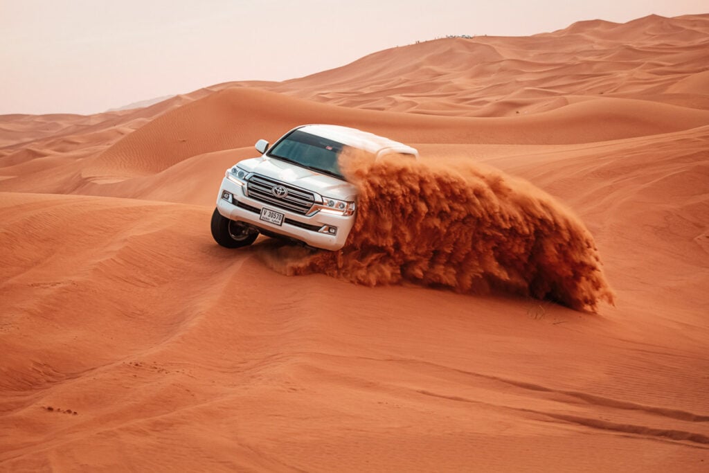 A white SUV drives quickly through orange sand dunes, kicking up a large spray of sand behind its tires.
