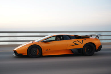 An orange sports car with SV logos drives on a road next to a body of water, with a person visible inside and a blurred background indicating motion.