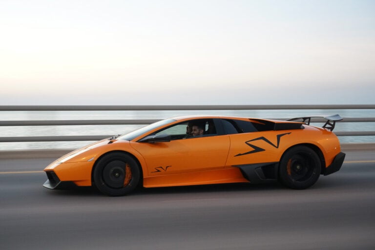 An orange sports car with SV logos drives on a road next to a body of water, with a person visible inside and a blurred background indicating motion.