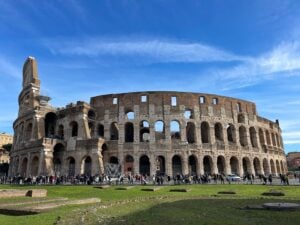 The Colosseum in Rome under a blue sky, with crowds of people walking along its perimeter and green grass in the foreground.