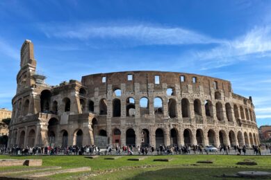 The Colosseum in Rome under a blue sky, with crowds of people walking along its perimeter and green grass in the foreground.