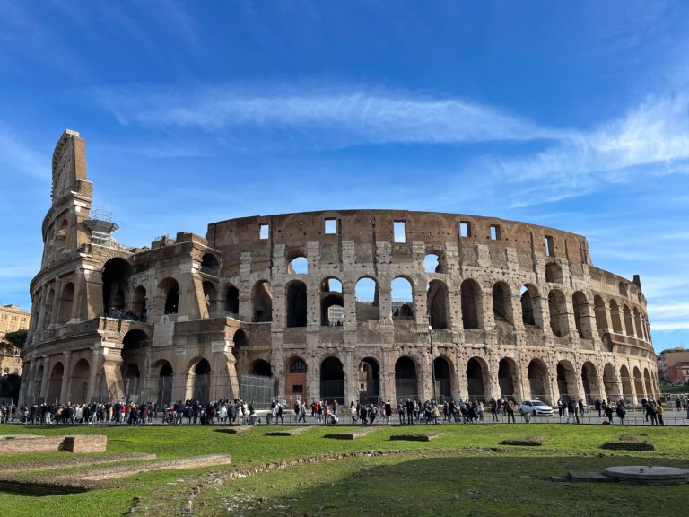 The Colosseum in Rome under a blue sky, with crowds of people walking along its perimeter and green grass in the foreground.
