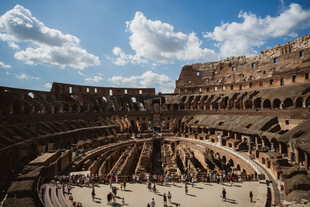A wide view of the interior of the Colosseum in Rome, showing its ancient stone structure, tiers, and tourists walking on the arena floor under a partly cloudy sky.