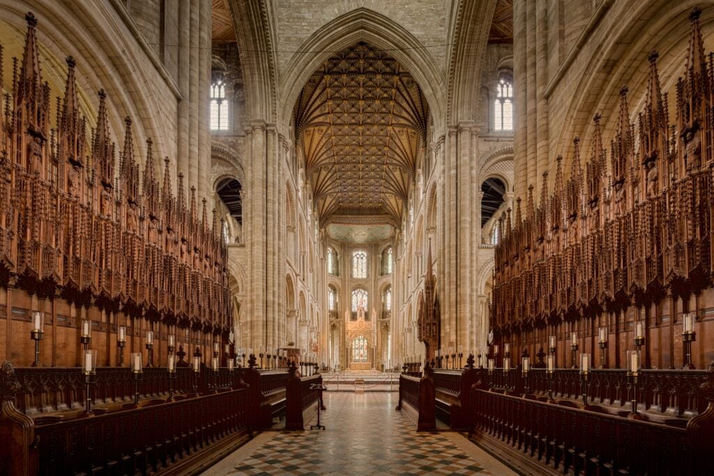 Interior view of a cathedral showing tall stone arches, intricate wooden choir stalls on both sides, and an ornate altar at the far end.